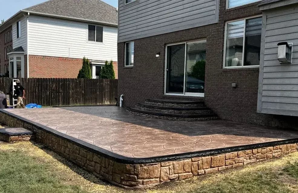 Raised stamped concrete patio with a stone-look border wall behind a brick home
