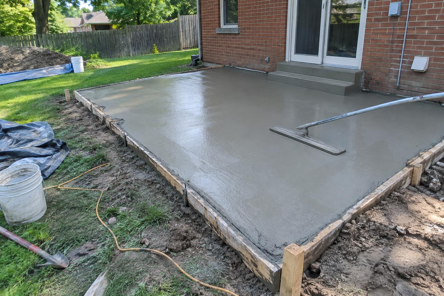 Fresh grey concrete patio slab being bull-floated inside wood forms behind a Plymouth home, lawn and fence in the background.
